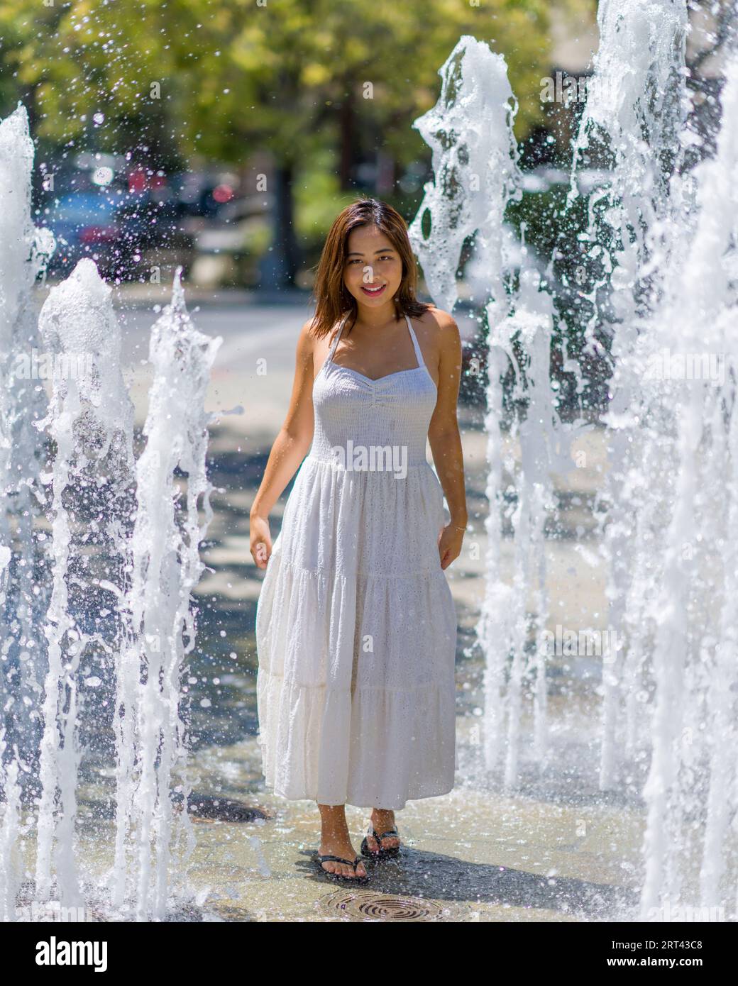 Belle jeune femme asiatique heureuse jouant dans la fontaine extérieure sur une journée chaude ensoleillée | White Sundress Banque D'Images