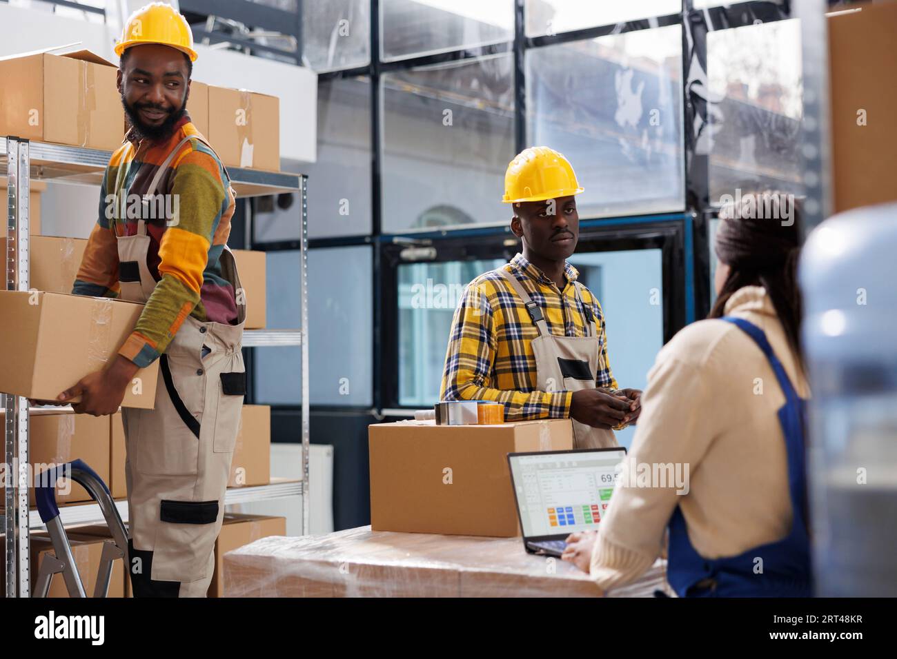 Manipulateurs de colis d'entrepôt et préparateur de commande travaillant dans la salle de stockage. Les employés des entrepôts portant des casques de sécurité prennent les boîtes en carton de l'étagère pendant que le responsable vérifie la liste des produits Banque D'Images