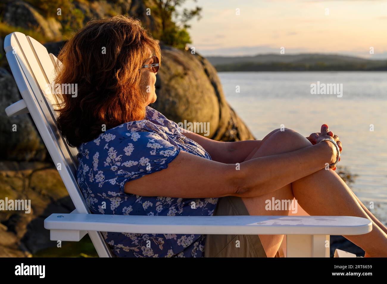 Une femme d'âge moyen de décence indienne est assise sur une chaise Adirondack en bois sur une jetée face au soleil couchant dans l'archipel de Kragero, Telemark, Norvège. Banque D'Images