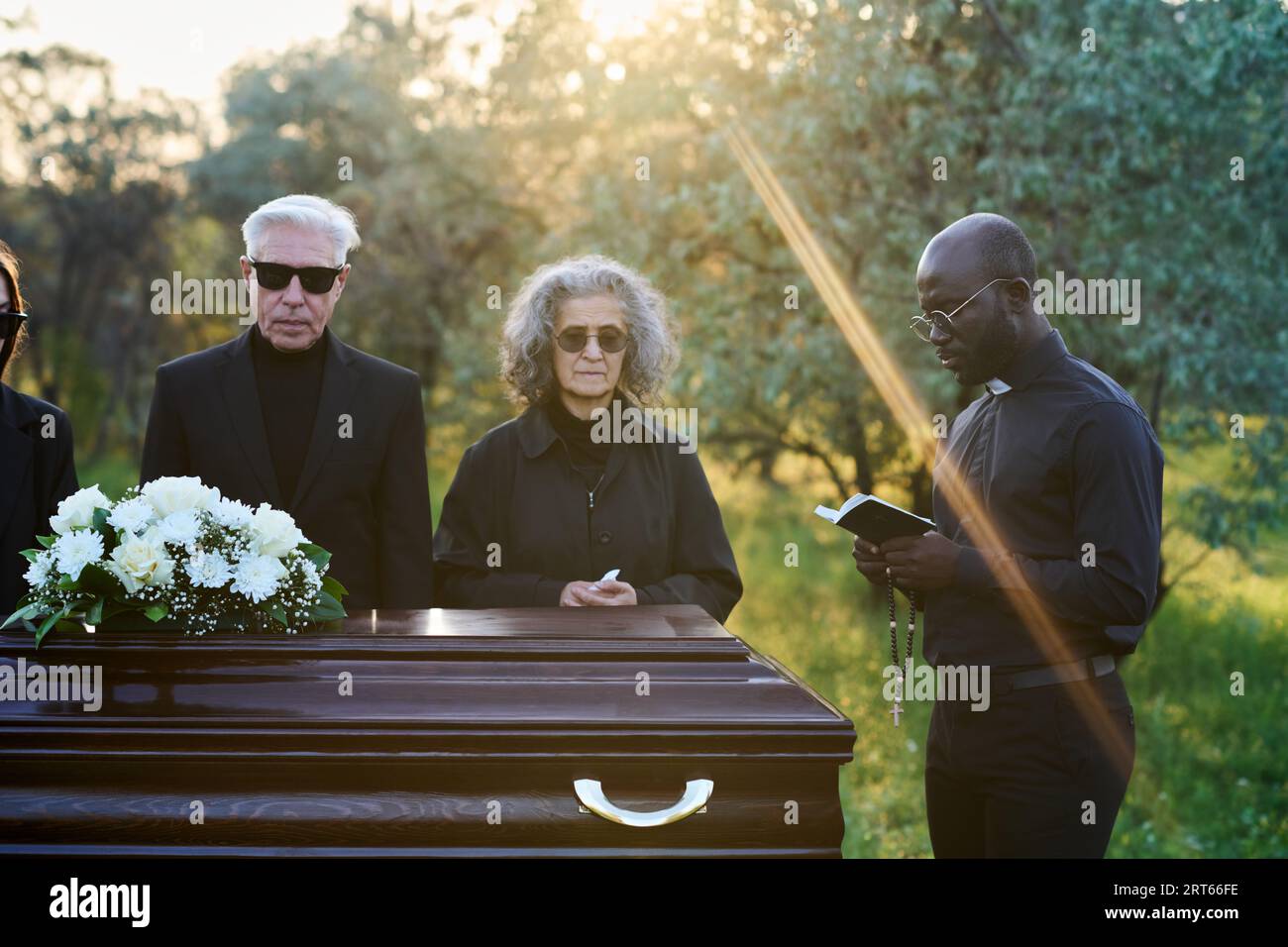 Jeune pasteur lisant des versets de la Sainte Bible tout en se tenant devant un cercueil avec le couvercle fermé et les parents en deuil de la personne décédée Banque D'Images