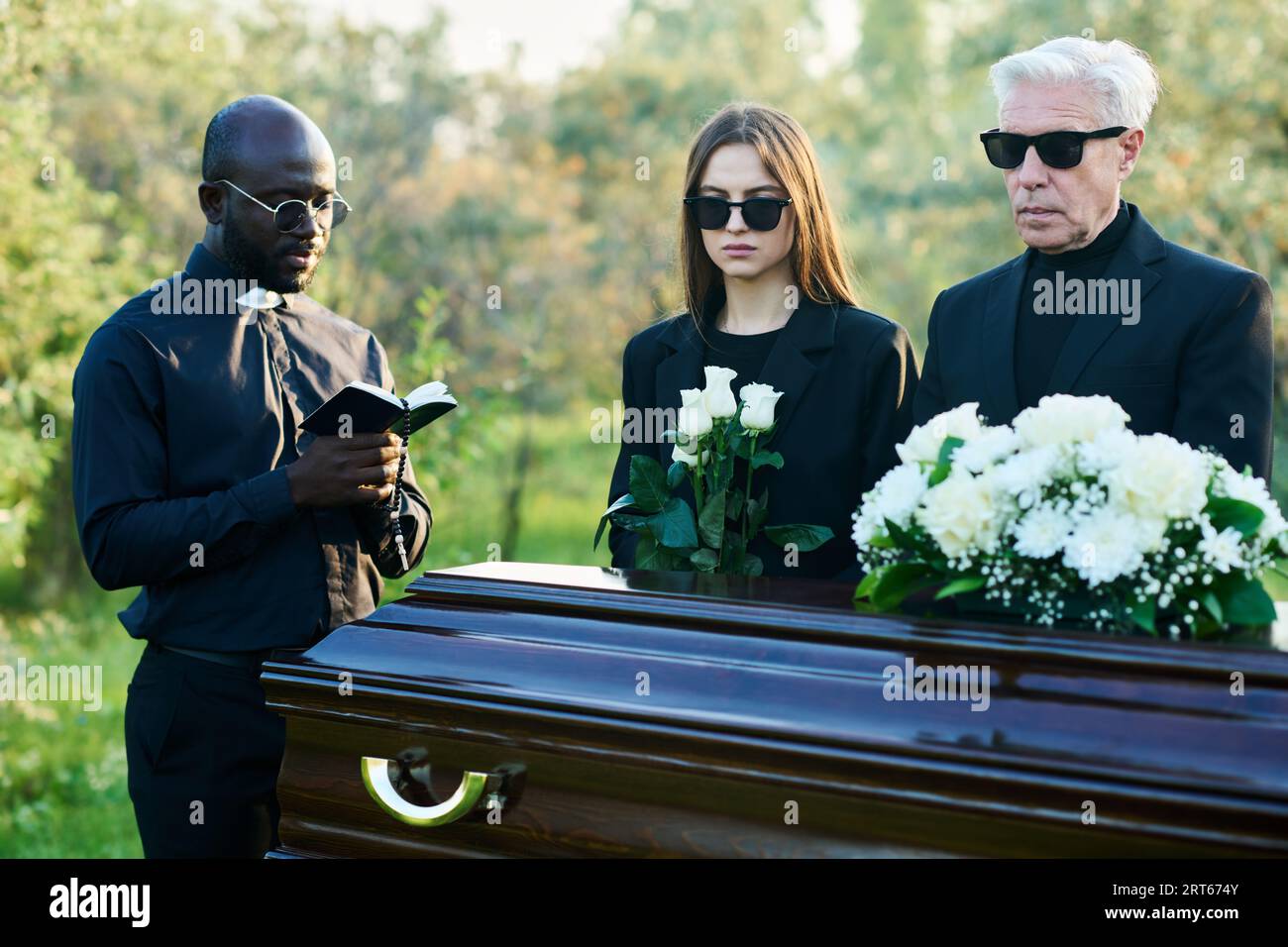 Homme mûr et jeune femme en vêtements de deuil et lunettes de soleil debout par cercueil tandis que prêtre lisant des versets de la Sainte Bible aux funérailles Banque D'Images