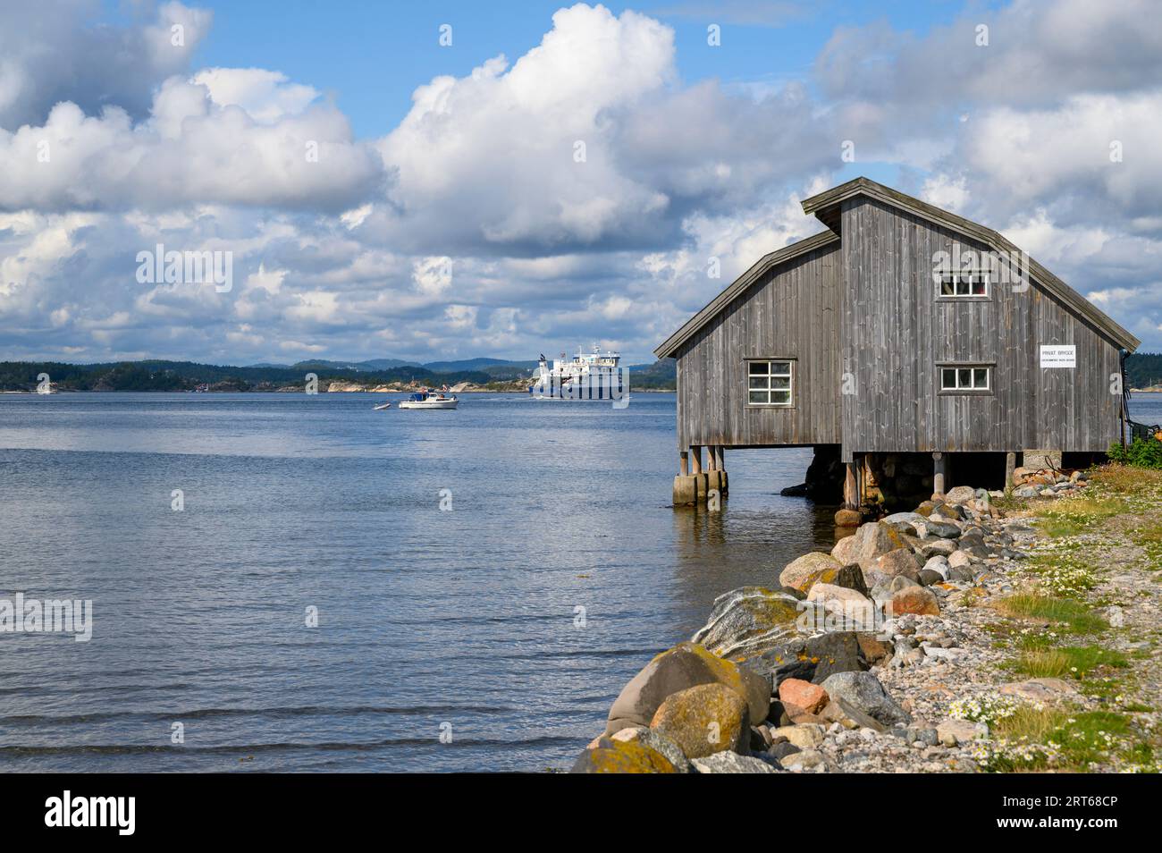 Un ferry pour voitures et passagers et un bateau de plaisance naviguent loin du port de l'île de Jomfruland devant un hangar à bateaux en bois patiné. Telemark, Norvège. Banque D'Images