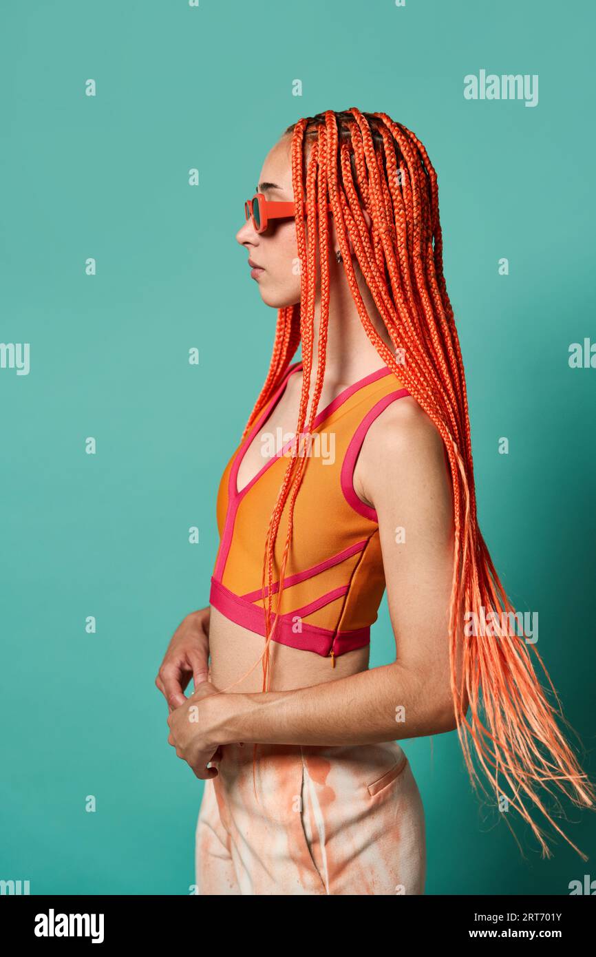 Vue latérale de jeune femme confiante avec de longues tresses afro orange portant des lunettes de soleil et un haut orange regardant loin sur fond turquoise Banque D'Images