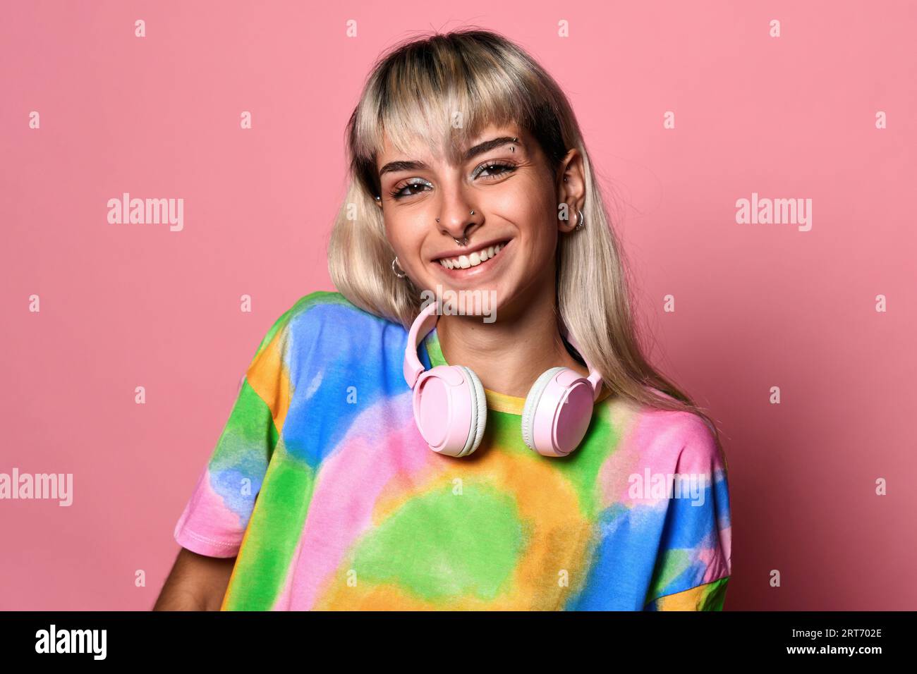 Jeune femme positive avec des piercings de visage et des cheveux blonds et des écouteurs autour du cou dans un t-shirt coloré debout sur fond rose Banque D'Images