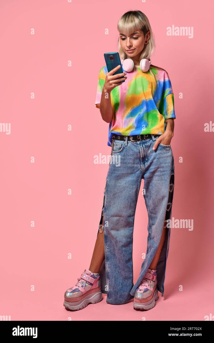 Corps complet de jeune femme en t-shirt coloré et jeans avec écouteurs autour du cou debout à l'aide d'un téléphone portable sur fond rose Banque D'Images