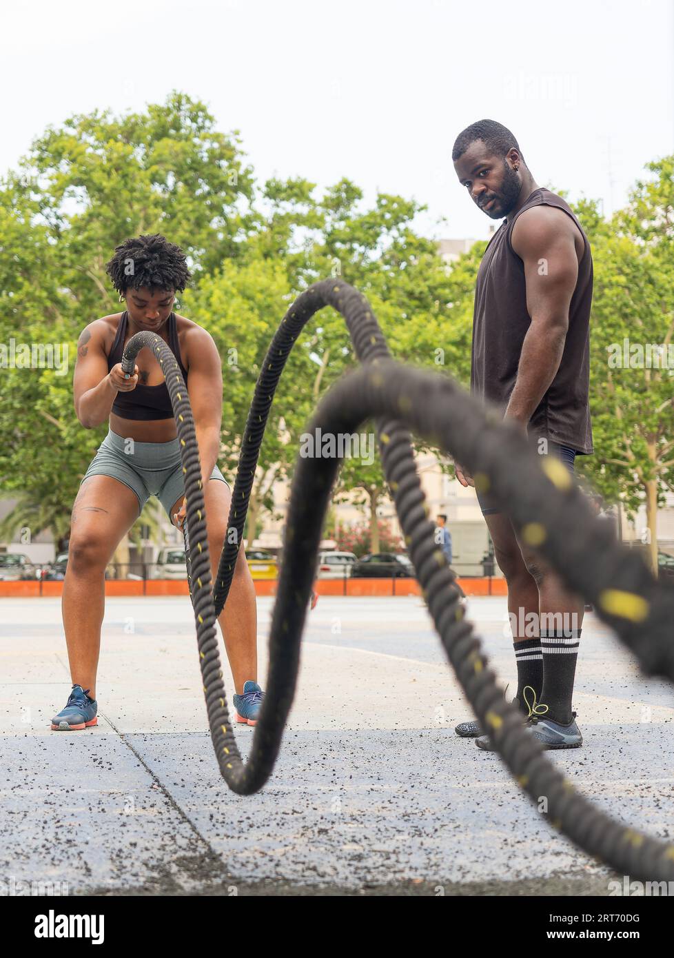 Athlète féminine afro-américaine forte s'exerçant avec des cordes de combat sur un terrain de sport avec instructeur masculin personnel pendant l'entraînement Banque D'Images