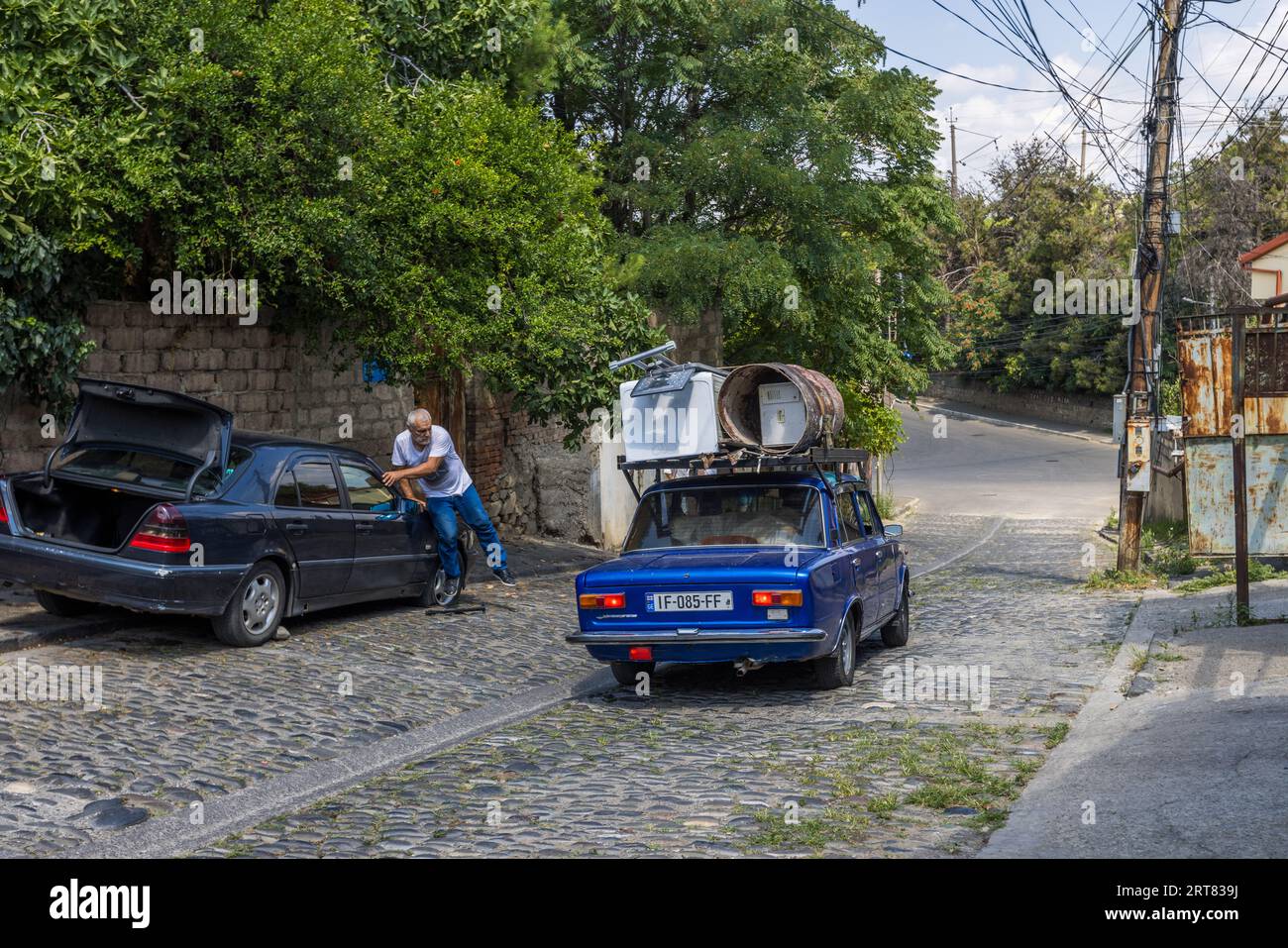 Un homme s'appuie contre la porte ouverte d'une voiture garée. Une voiture bleue avec une machine à laver et un tonneau sur le toit est garée dans la rue pavée. Tbilissi, Chugureti, Géorgie Banque D'Images