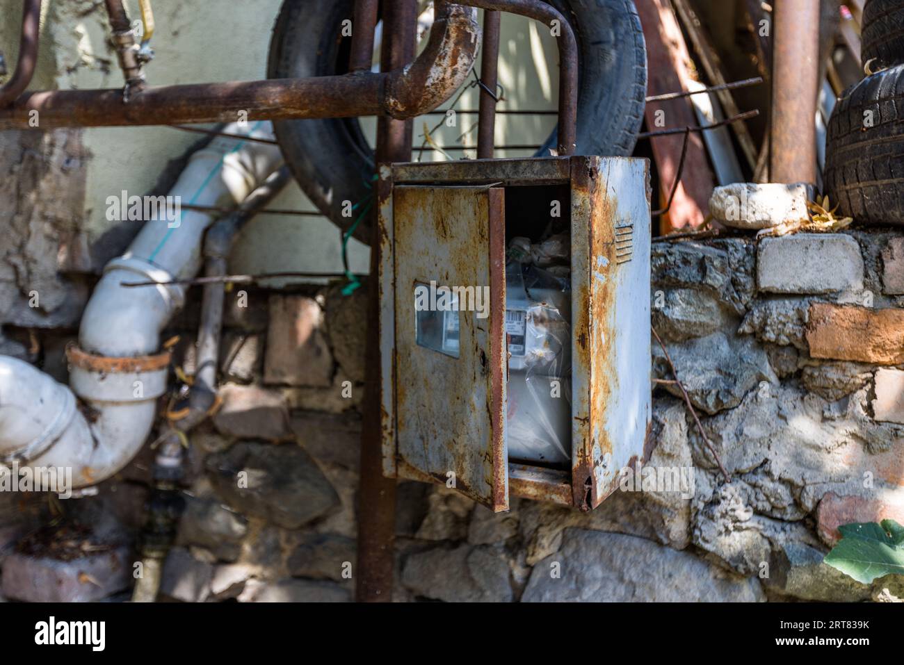 Un boîtier de compteur électrique rouillé avec une porte ouverte est accroché à un mur en pierre naturelle dans le quartier de Chugureti à Tbilissi, en Géorgie. Des tuyaux rouillés, un tuyau d'évacuation blanc et de vieux pneus l'entourent. Le compteur est visible à l'intérieur du boîtier derrière un bâche en plastique. Banque D'Images