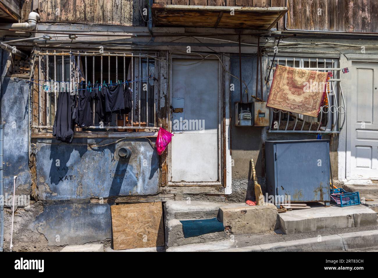 Le linge sèche devant une fenêtre grillagée sur la façade d'une vieille maison de la rue Kumisi dans le quartier de Chugureti à Tbilissi, en Géorgie. Un tapis à motifs est suspendu pour aérer au-dessus de la grille de la fenêtre voisine. Des tuyaux et un boîtier de compteur électrique sont fixés à même le mur. Un balai en brindilles est posé contre la porte à côté d'une armoire métallique. Banque D'Images
