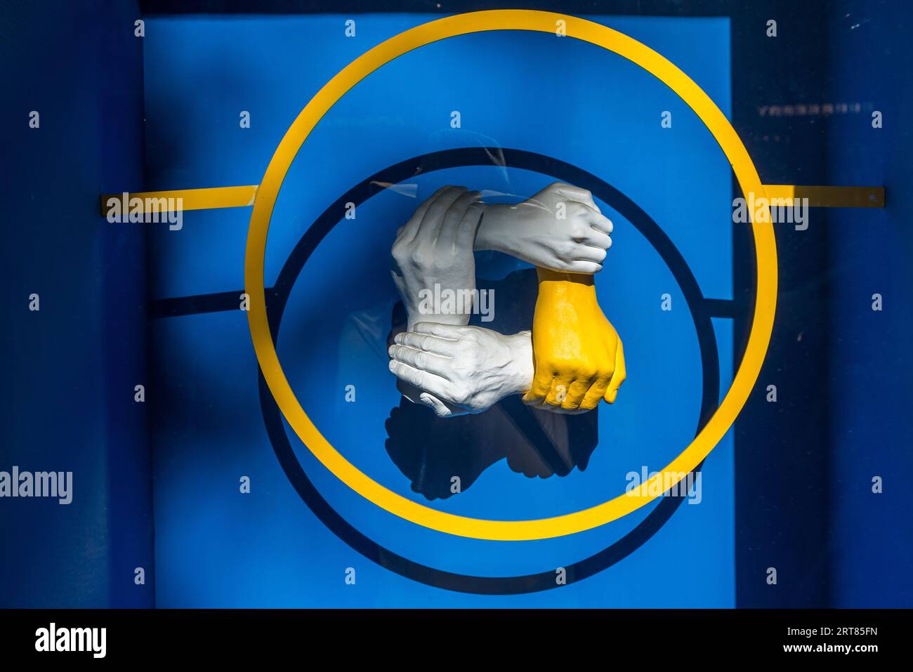 Une sculpture de quatre mains entrelacées se trouve dans une vitrine à Tbilissi, en Géorgie (peut-être au Centre d'information sur l'OTAN et l'UE sur la Place de la Liberté). Trois mains sont blanches, une est jaune. Chaque main saisit le poignet de la suivante. Les mains reposent sur un fond bleu à l'intérieur d'un anneau jaune. Une barre jaune rencontre l'anneau à gauche et à droite. Banque D'Images