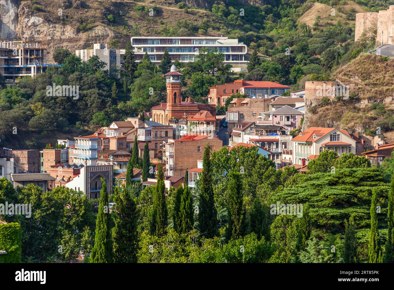 Le quartier des bains d'Abanotubani (géorgien : აბანოთუბანი) baigné par la lumière du matin sur le flanc en contrebas de la forteresse de Narikala dans la vieille ville de Tbilissi, en Géorgie. Le minaret en brique de la mosquée Juma s'élève parmi les maisons aux toits de tuiles rouges. En bas à gauche, un arc pointu carrelé s'ouvre, probablement la façade du bain Orbeliani (Chreli Abano, "bain coloré"). De petites dômes bleus se courbent à côté. En haut à droite, les murs et une tour de la forteresse se dressent sur le rocher. Un long bâtiment moderne et une coquille de bâtiment se trouvent plus haut sur le flanc. Des cyprès et des arbres caduques poussent au premier plan. La ville n Banque D'Images