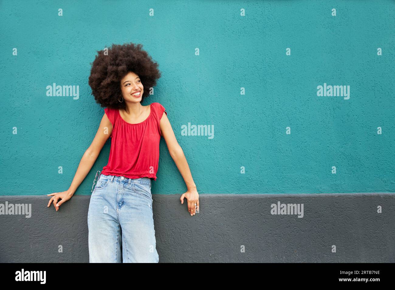 Souriant mignon jeune femme marocaine avec coiffure afro regardant loin tout en se tenant debout appuyé sur le mur bleu avec les mains sur la surface grise dans la lumière du jour Banque D'Images