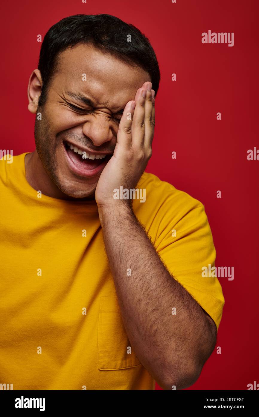 expression du visage, homme indien embarrassé en t-shirt jaune riant et touchant le visage sur fond rouge Banque D'Images