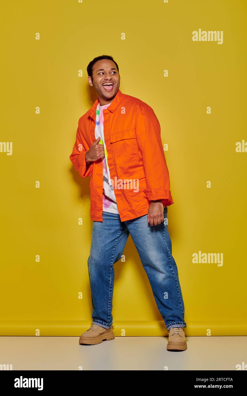 pleine longueur d'homme indien excité en veste orange et jeans en denim debout sur fond jaune Banque D'Images