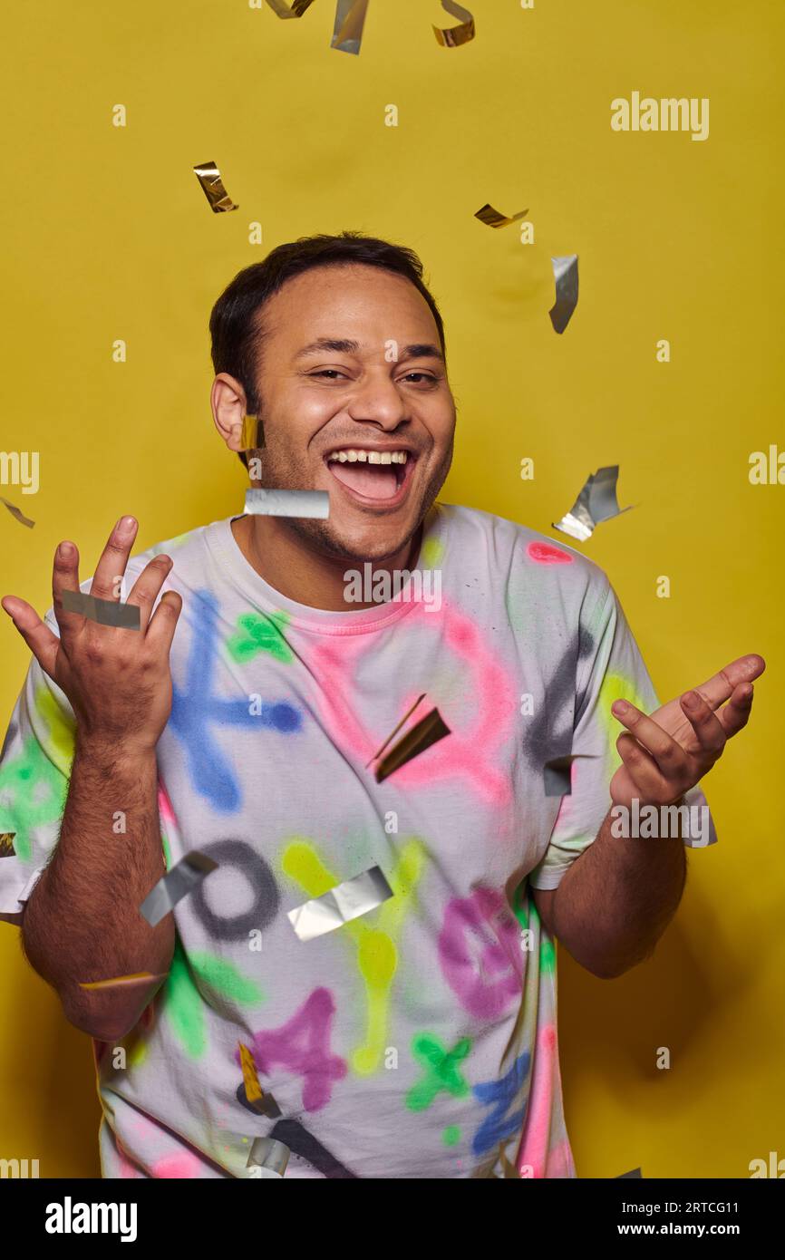 homme indien positif en t-shirt faisant des gestes près de la chute de confettis sur fond jaune, concept de fête Banque D'Images