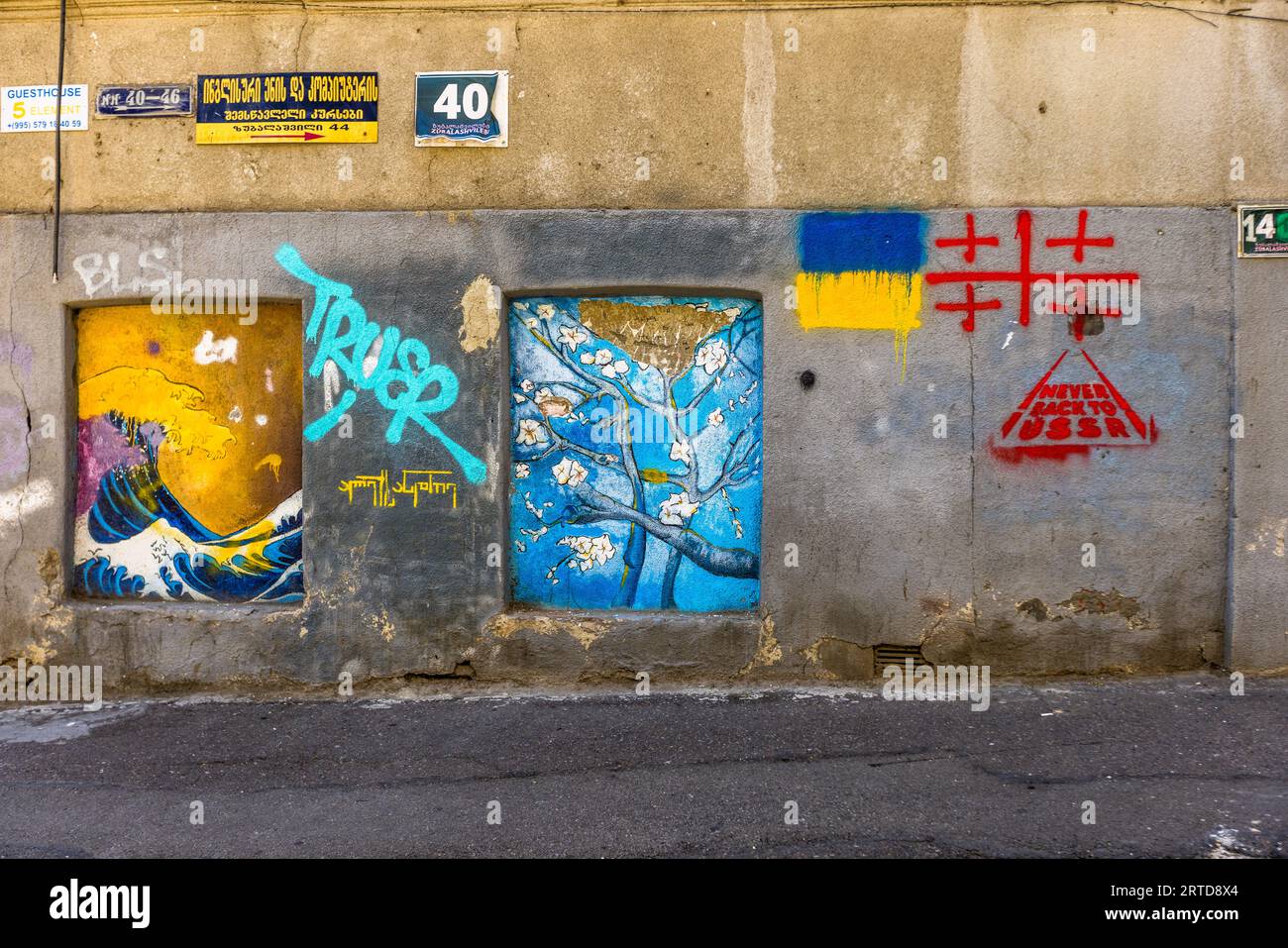 Un mur à Tbilissi, en Géorgie, est couvert de graffitis et de peintures murales. Sur la gauche se trouve une vague contre un ciel jaune. Au centre se trouve un arbre avec des fleurs blanches sur fond bleu. Sur la droite se trouve un drapeau ukrainien et les mots "NE JAMAIS RETOURNER À l'URSS". Au sommet se trouvent des panneaux de signalisation Banque D'Images