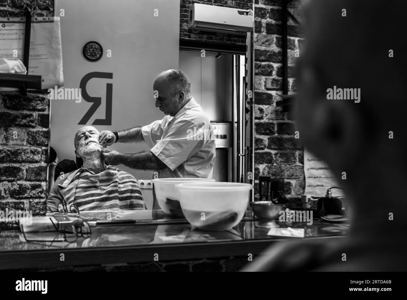 Un coiffeur se penche sur un client âgé dans un salon de coiffure de la rue Pushkin dans la vieille ville de Tbilissi, en Géorgie. Il tient le visage savonné du client avec ses mains. Le client est allongé, les yeux fermés, un cape rayée sur les épaules. La scène apparaît dans le miroir du salon, ce qui explique pourquoi le logo "RL" sur le mur est inversé. Deux bols blancs sont posés sur une étagère en verre devant le miroir. Un climatiseur est accroché au mur en briques. Banque D'Images