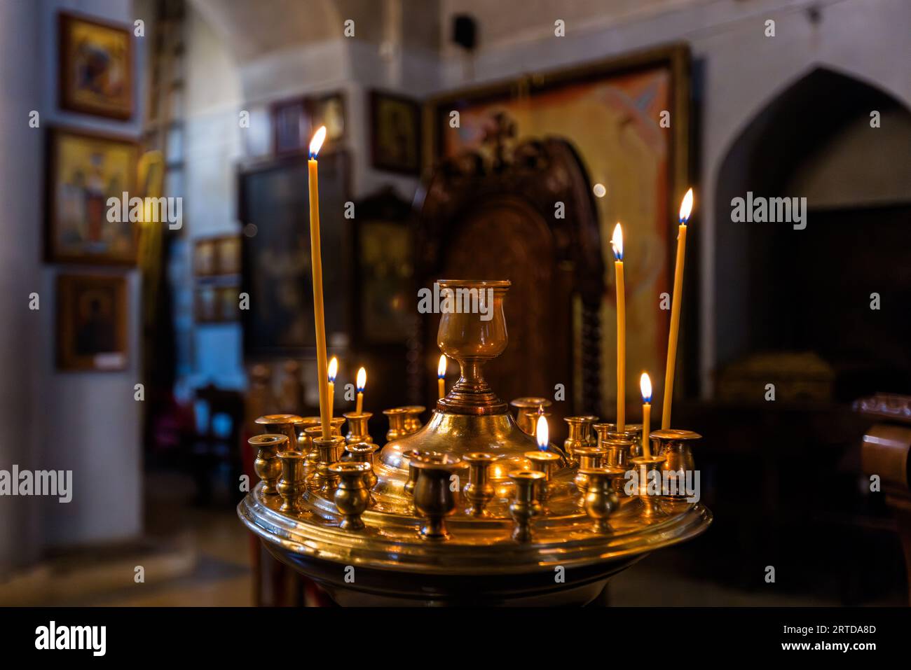 De fines bougies de cire brûlent sur un chandelier rond en laiton à l'intérieur de l'église Metekhi dans le quartier d'Avlabari à Tbilissi, en Géorgie. Le chandelier possède de petites prises tout autour et un support en forme de calice au centre. Des icônes sont accrochées à des murs simples et clairs en arrière-plan, ainsi qu'une chaise en bois sculpté à haut dossier et une niche en arc pointu. Le roi Démétrius II fit construire l'église actuelle de l'Assomption vers 1278 à 1284. Elle se dresse sur une falaise abrupte au-dessus de la rivière Mtkvari (Koura). Le roi Vakhtang Ier Gorgasali, considéré comme le fondateur de la ville, y construisit une église et une forteresse comme résidence. Banque D'Images