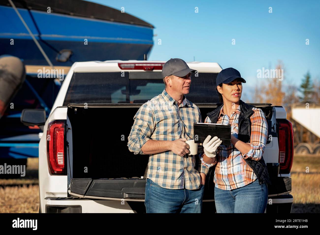 Mari et femme prenant un café tout en se penchant sur le hayon de leur camion d'une demi-tonne et en utilisant un appareil portable sans fil pour gérer et surveiller t... Banque D'Images