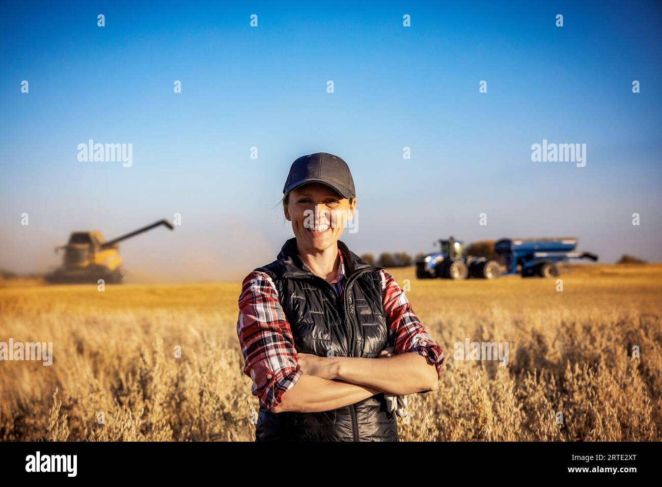 Portrait d'une fermière mature debout dans un champ de céréales, posant et souriant pour la caméra pendant la récolte avec une moissonneuse-batteuse se préparant à décharger... Banque D'Images