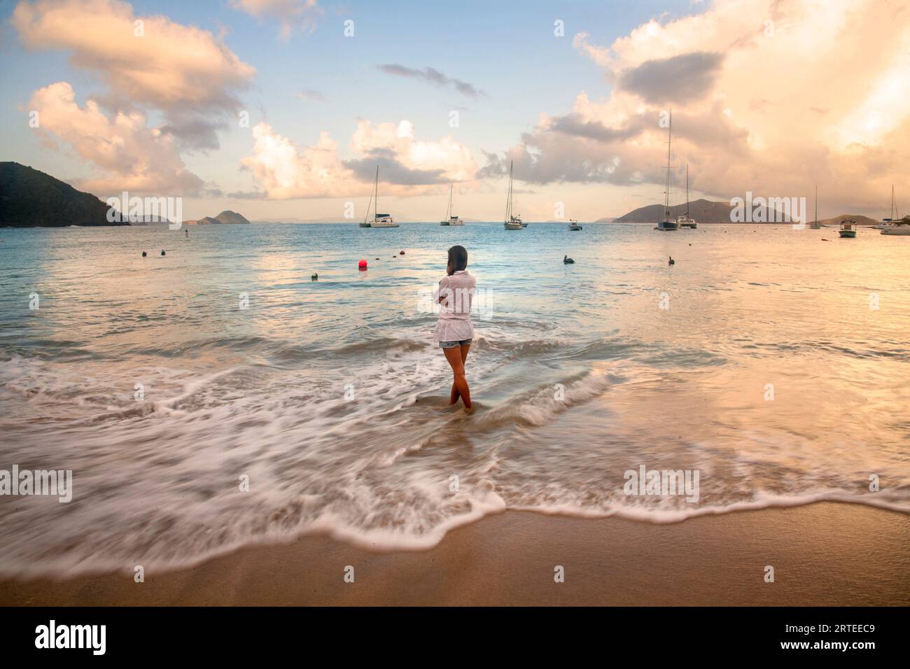 Vue prise de derrière d'une femme debout dans le surf mousseux sur la plage, regardant l'eau turquoise avec des bateaux amarrés au large... Banque D'Images