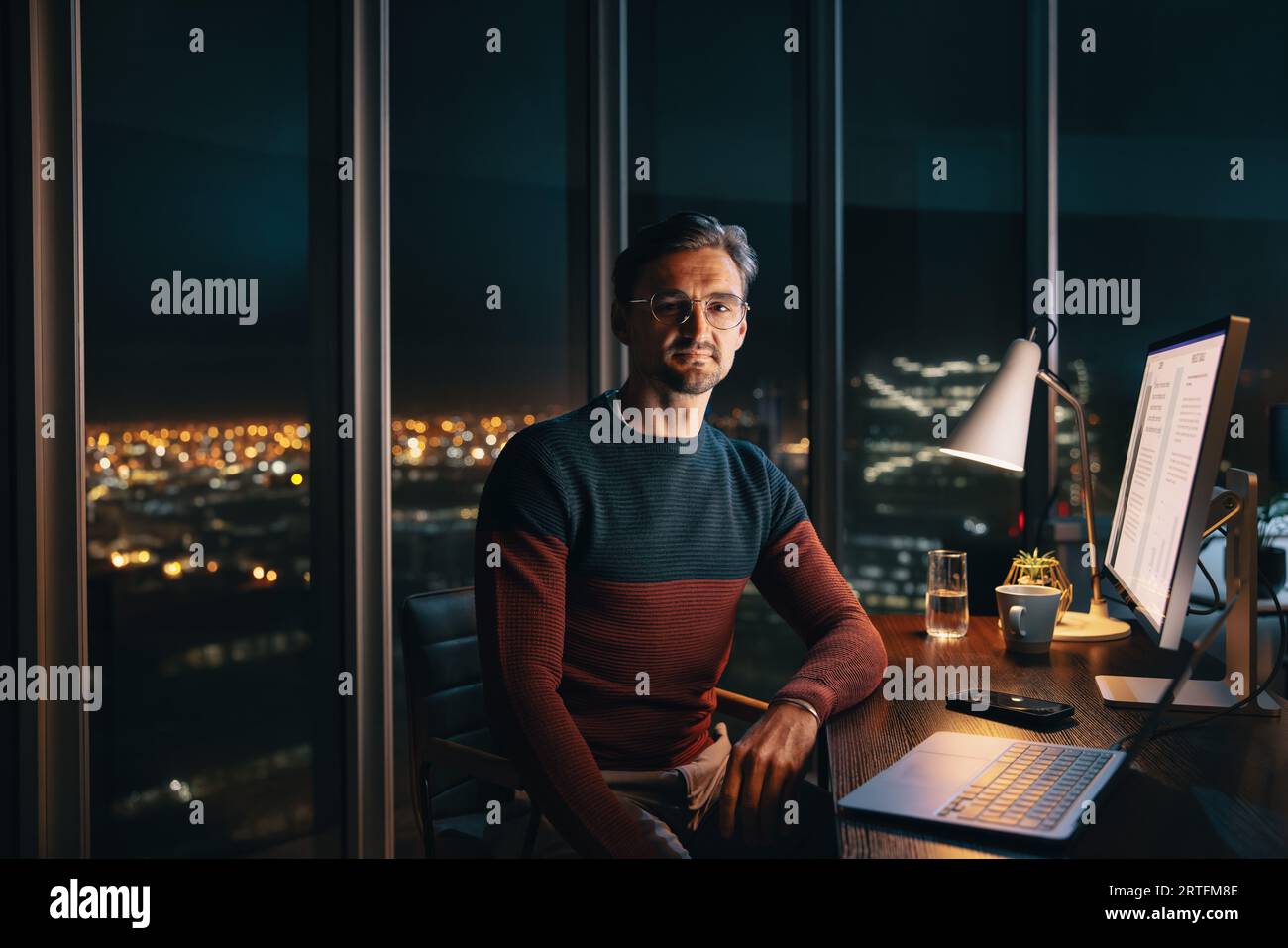 Un homme d'affaires caucasien concentré travaillant tard dans la nuit dans un bureau. Il est assis à son bureau, regardant la caméra tout en travaillant avec diligence sur un ordinateur portable. Banque D'Images