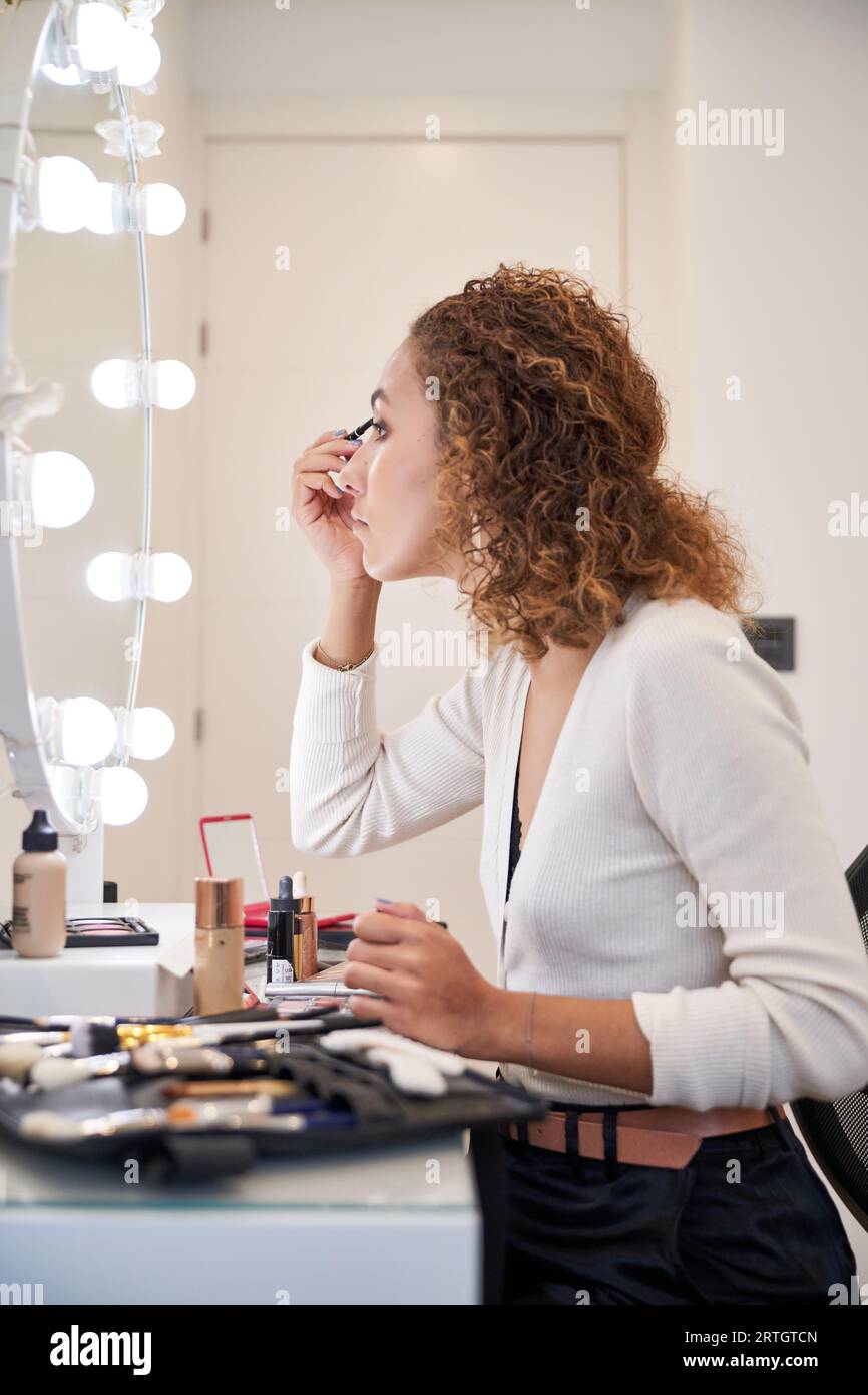 Vue latérale de jeune femme aux cheveux bouclés faisant le maquillage des yeux avec des produits cosmétiques assis devant le miroir éclairé Banque D'Images