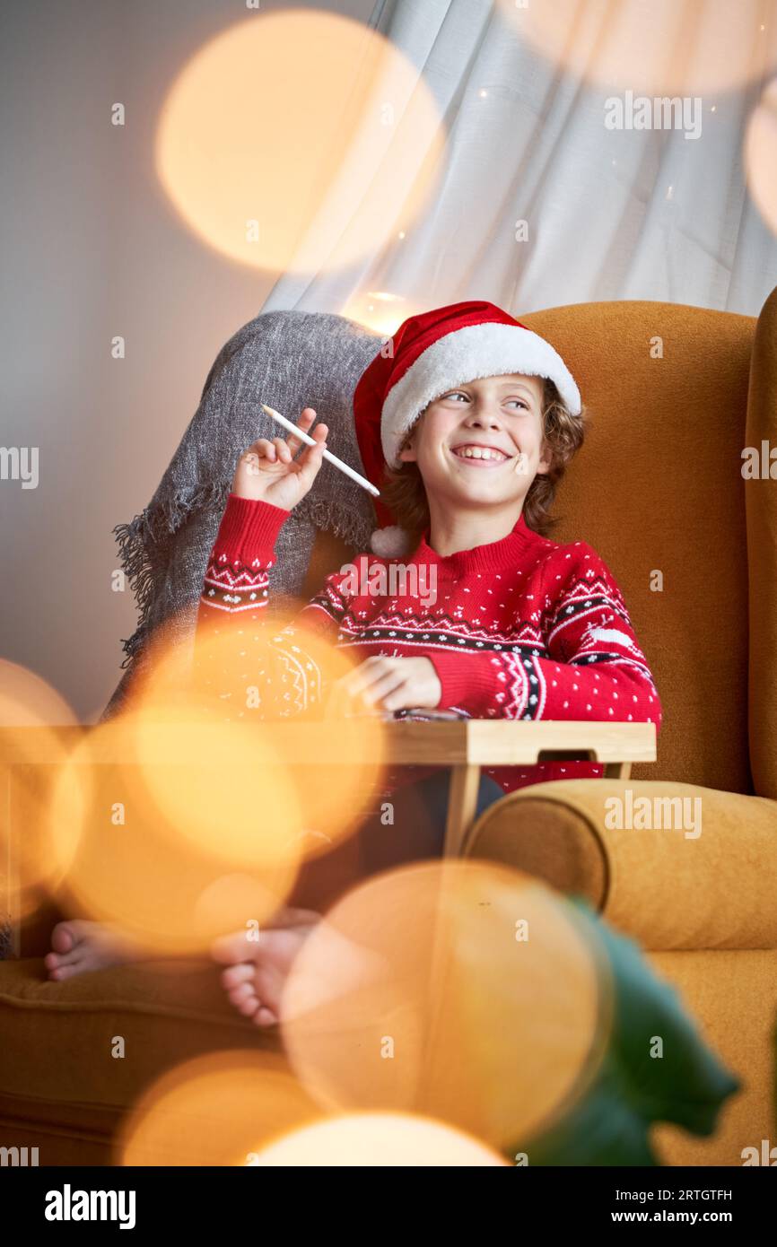 Petit enfant heureux en pull rouge et chapeau de Père Noël regardant loin tout en étant assis sur un fauteuil et dessiner avec un crayon Banque D'Images