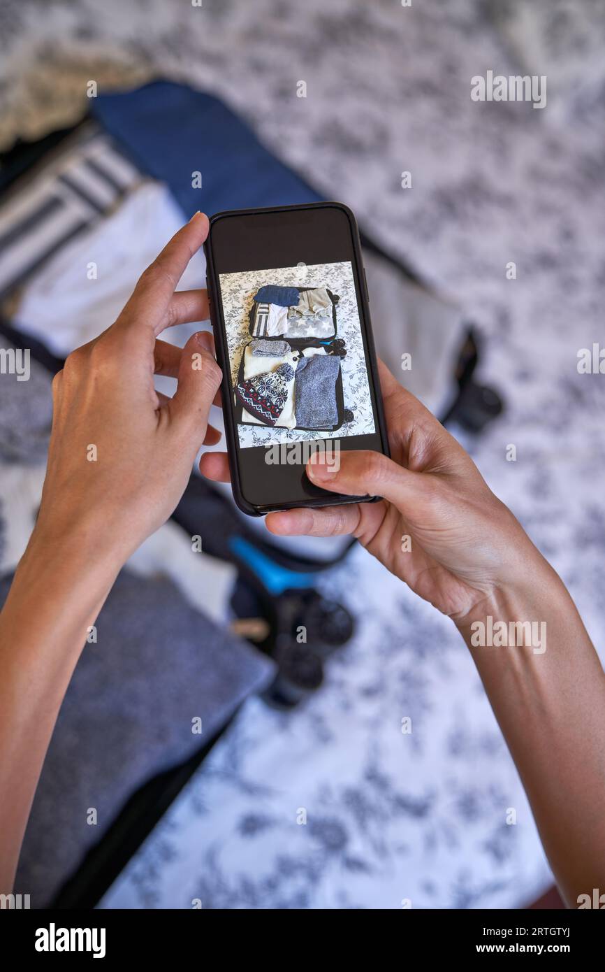 Mise au point sélective angle élevé de la personne anonyme sans visage prenant la photo de vêtements dans les bagages sur smartphone placé sur le lit Banque D'Images