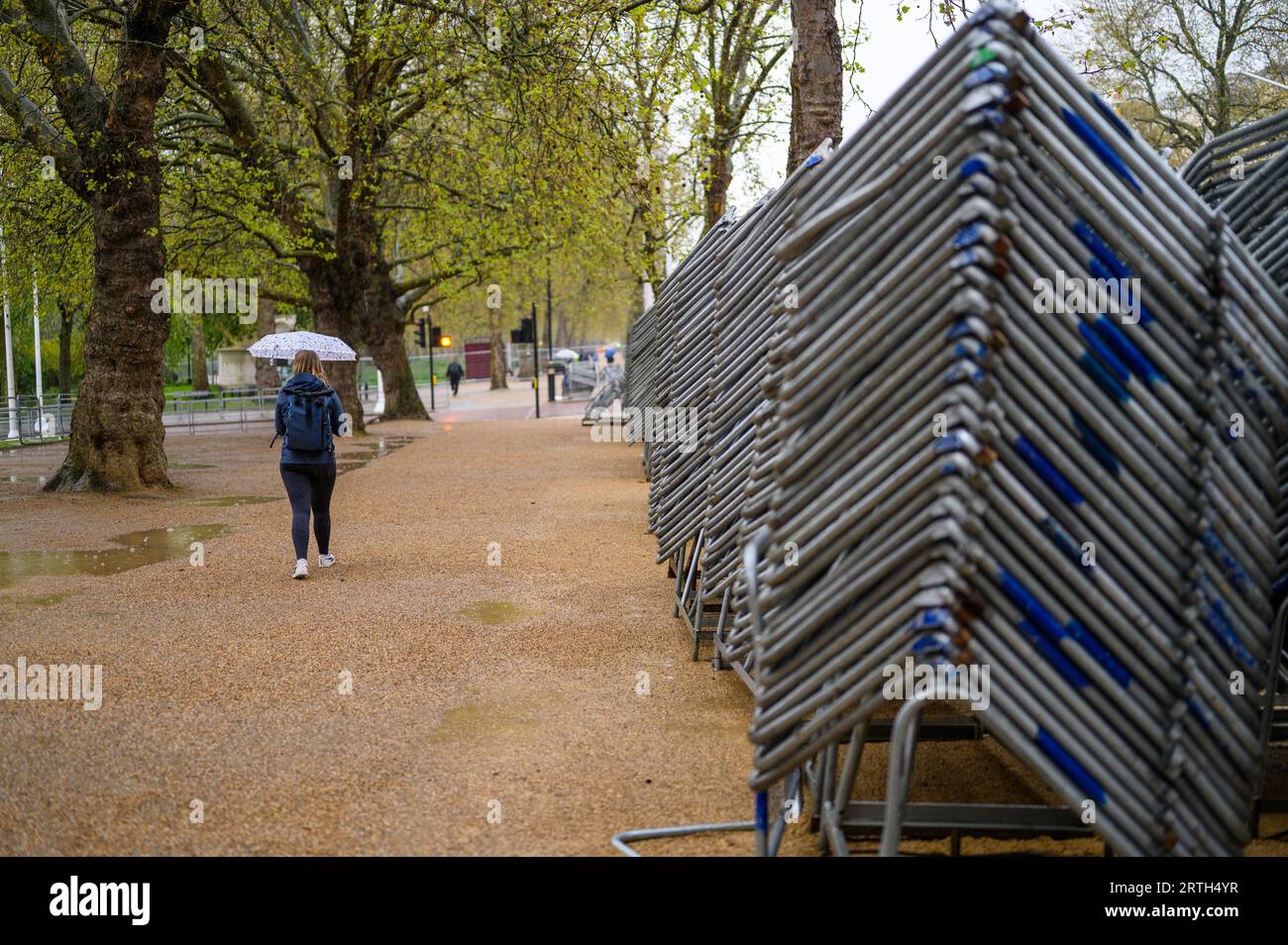LONDRES - 24 avril 2023 : •Une femme avec un parapluie et un sac à dos passe devant des barrières métalliques empilées haut sur le centre commercial, prête pour le couronnement du roi Charles Banque D'Images