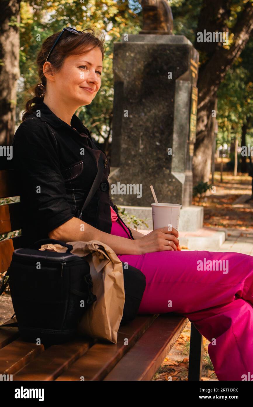 Fille avec tasse à café assise sur le banc dans le parc. Femme se détendant dans l'allée d'automne. Tombez au centre-ville. Style de vie urbain. Vacances par jour ensoleillé. Banque D'Images