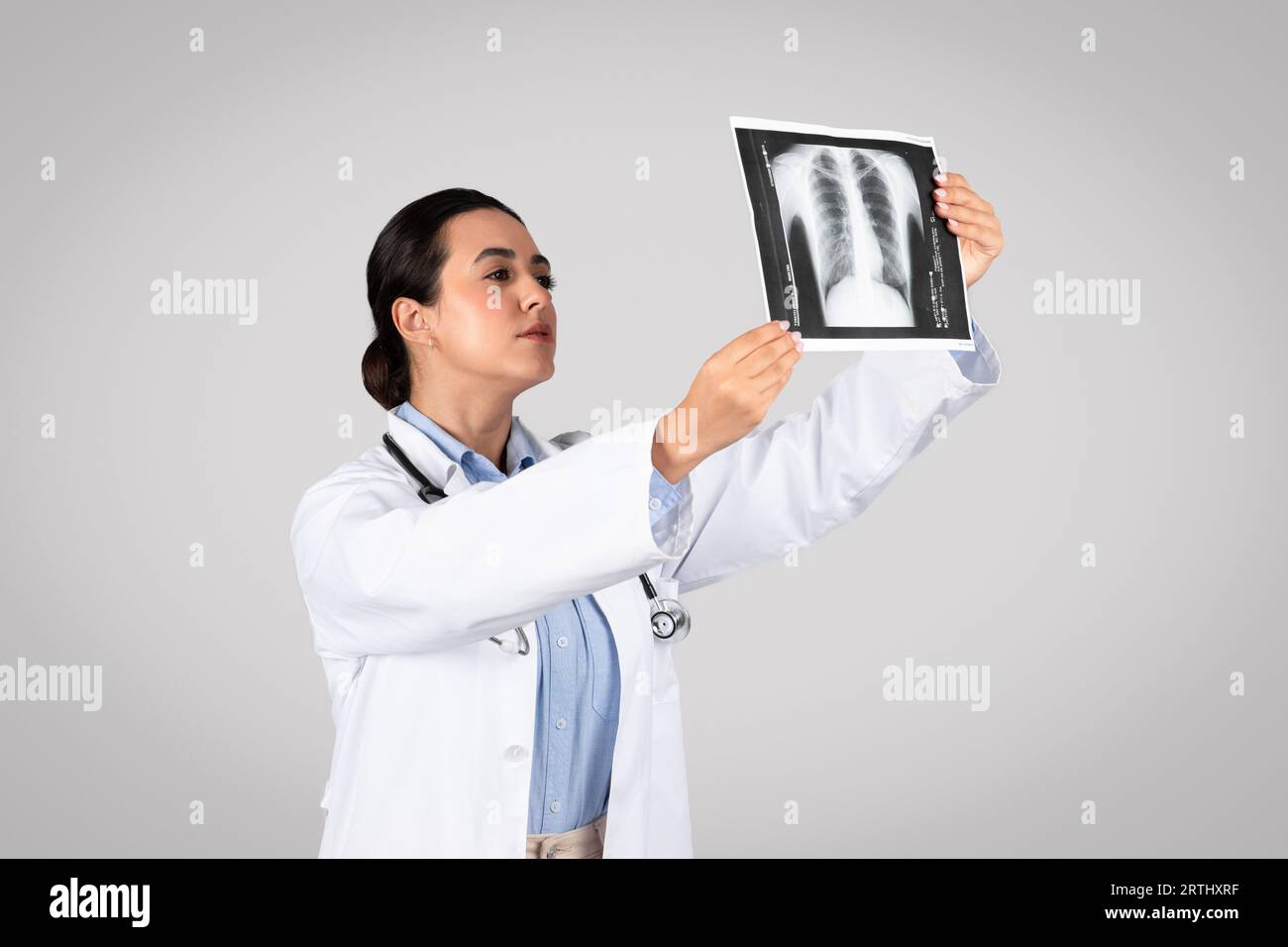 Jeune médecin latine tenant et regardant la radiographie thoracique, debout sur fond gris de studio Banque D'Images
