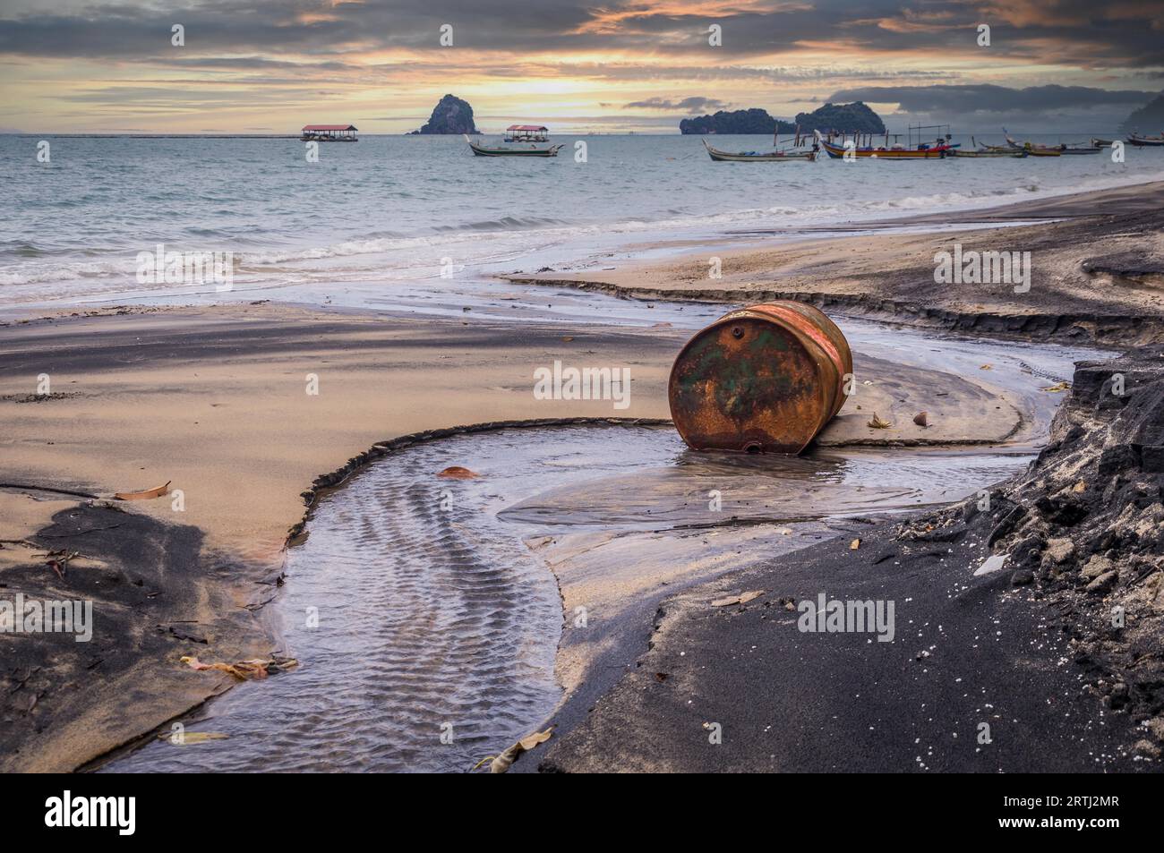 Le baril de pétrole rouillé sur une plage en partie noire illustre la pollution de l'environnement par les déversements de pétrole. Bateaux Longtail en arrière-plan au coucher du soleil Banque D'Images