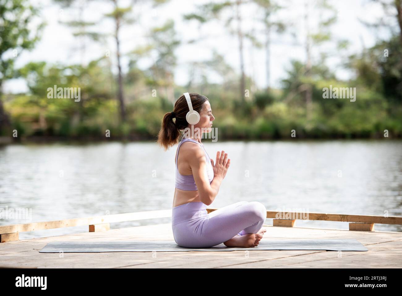 Femme sportive pratiquant le yoga sur une jetée au bord d'un lac, assise méditant avec des écouteurs, partie de la série. Banque D'Images