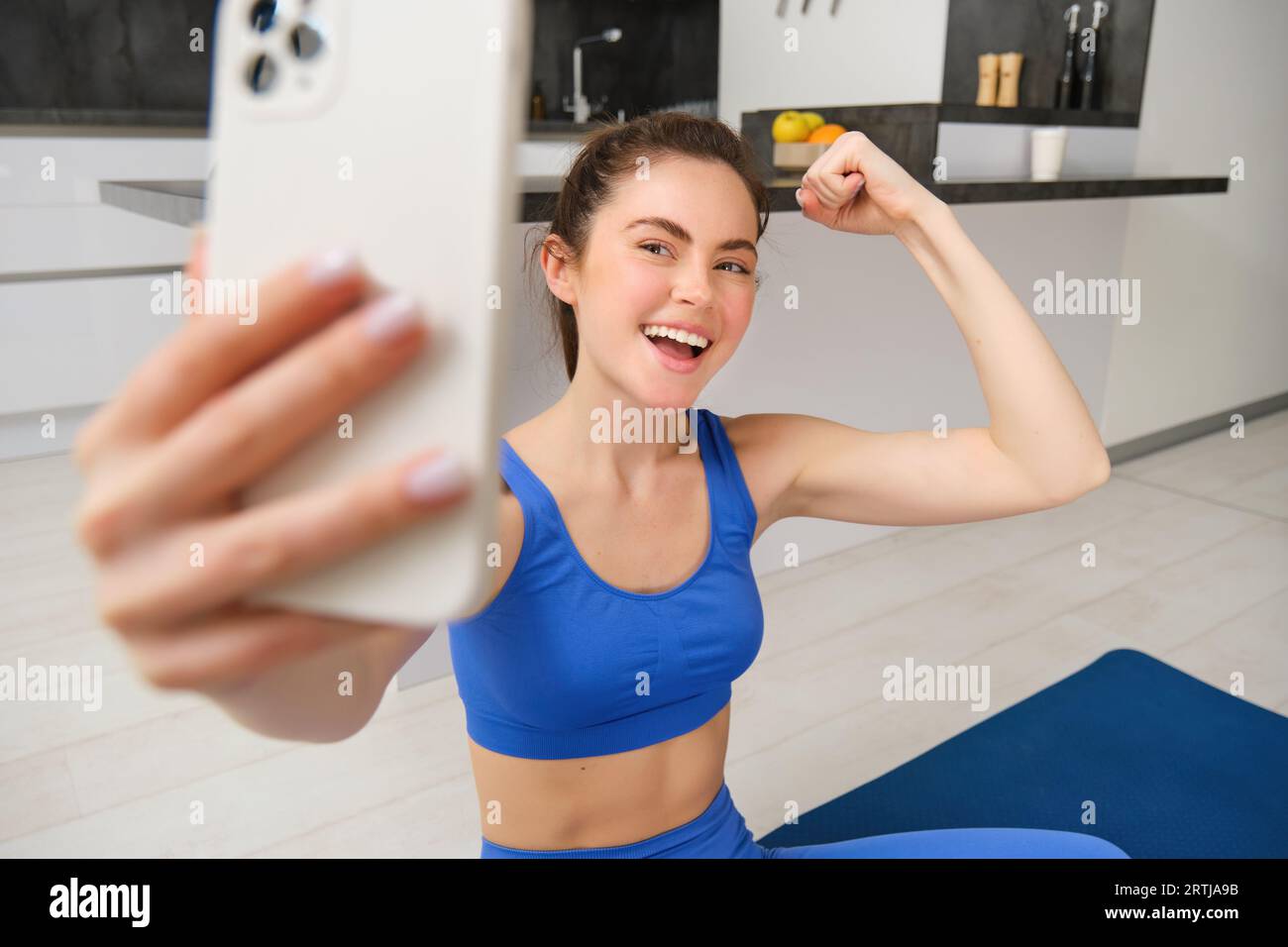 Portrait de fille de fitness active, prend selfie sur smartphone pendant l'entraînement à la maison, posant sur le tapis de fitness, montre ses muscles forts et rit Banque D'Images