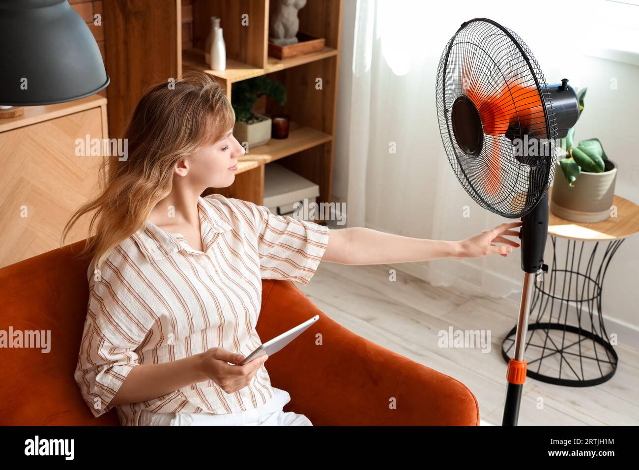 Jeune femme avec tablette et ventilateur électrique soufflant assis sur le canapé à la maison Banque D'Images