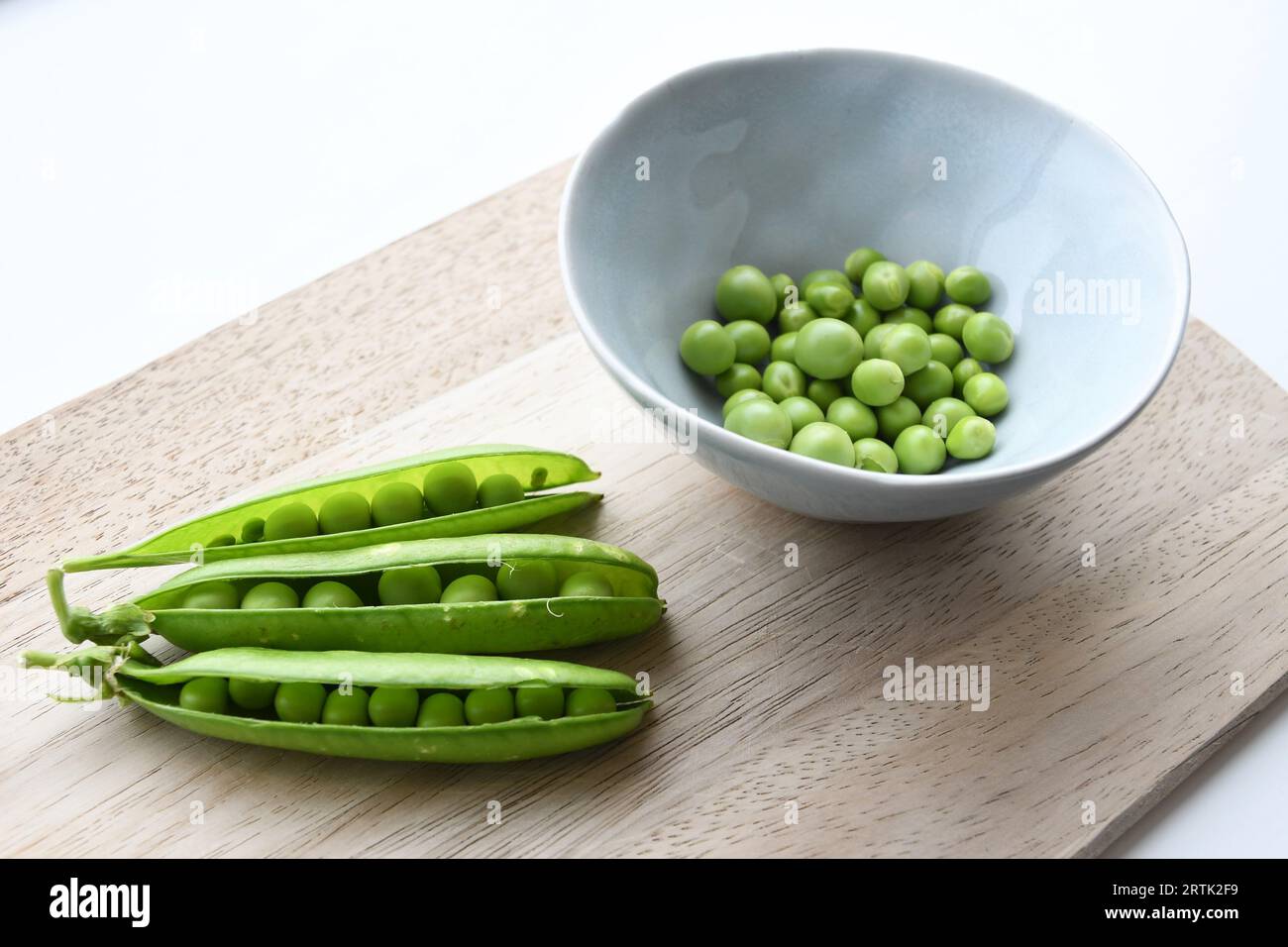 Pois du jardin (Pisum sativum) dans un bol en céramique bleu avec des gousses de pois, sur une planche à découper en bois. Isolé sur fond blanc. Orientation paysage. Banque D'Images