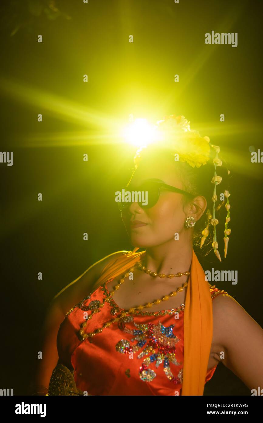 Une danseuse sundanaise pose devant une lumière brillante tout en portant un costume orange avec des lunettes de soleil et des fleurs sur la tête la nuit Banque D'Images