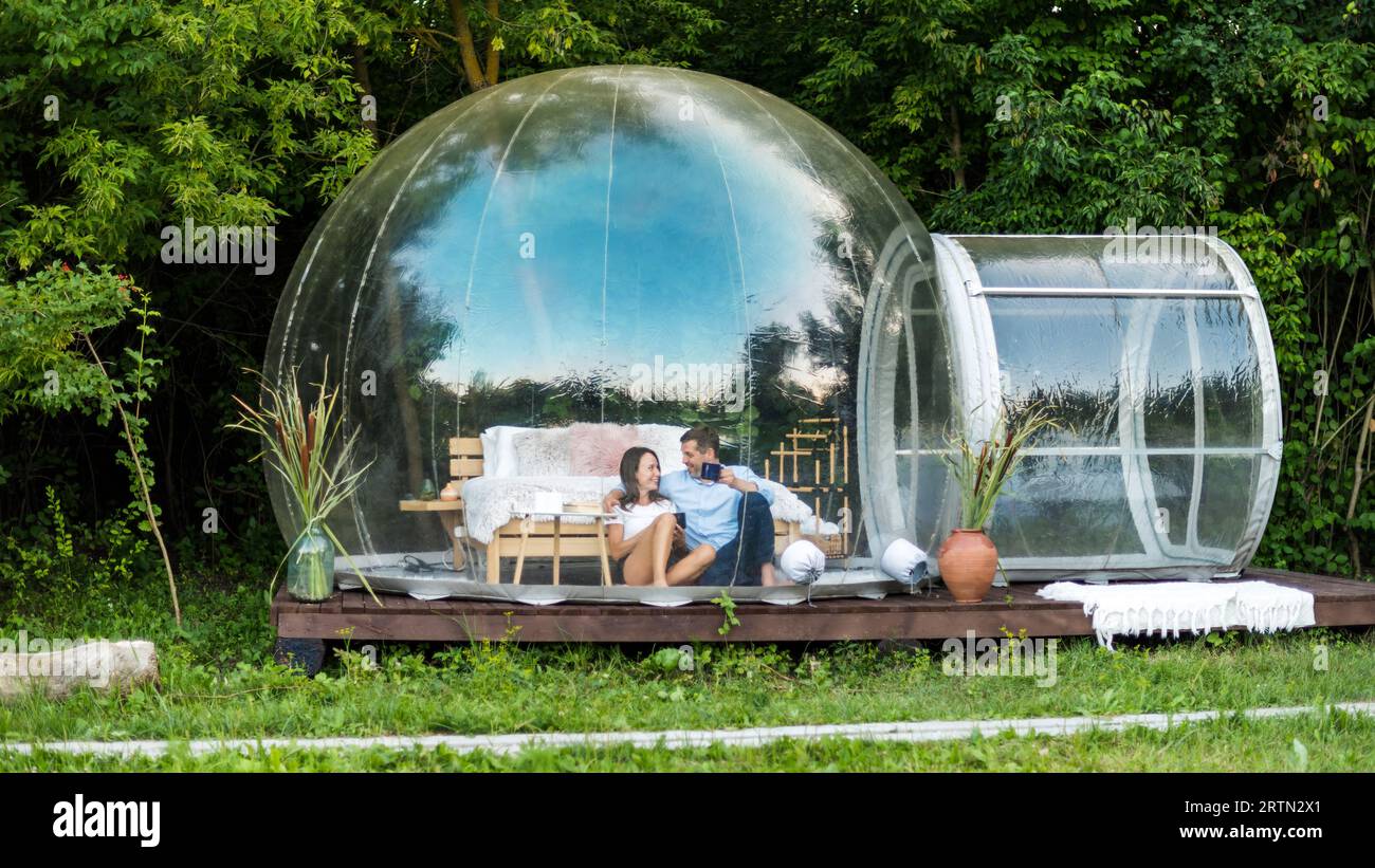 Un couple heureux assis à l'intérieur de tente bulle transparente au glamping avec des tasses, vue de l'extérieur. Forêt luxuriante sur le fond Banque D'Images