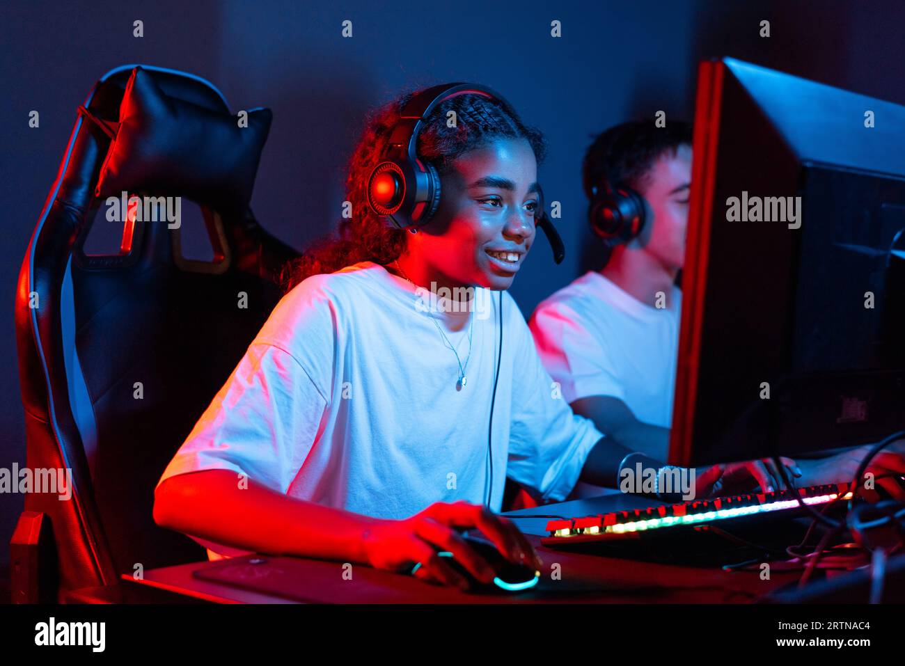 Garçon blanc et fille noire adolescents dans des casques jouant à des jeux vidéo dans un club de jeux vidéo avec éclairage bleu et rouge. Clavier et souris avec éclairage Banque D'Images