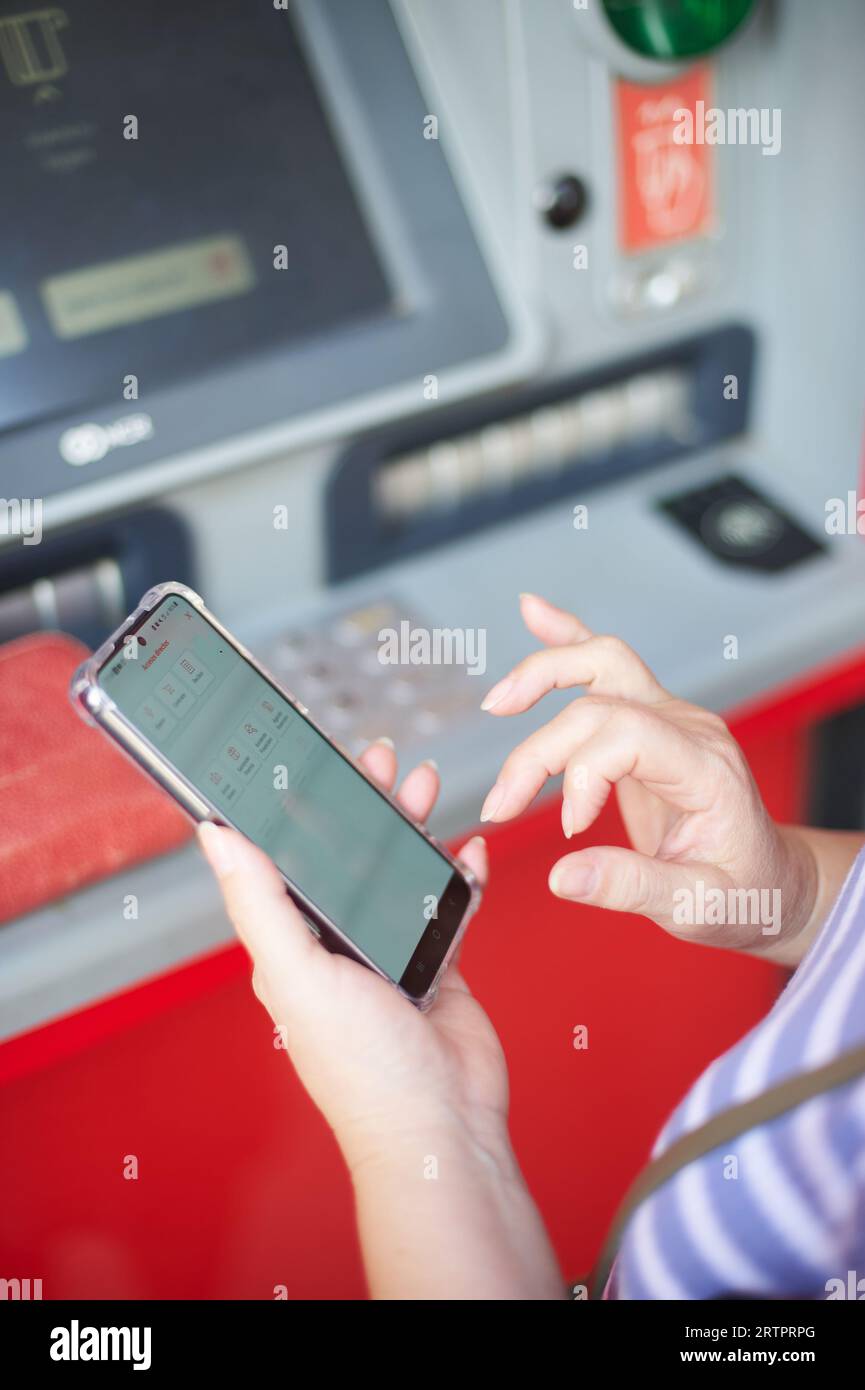 Femme méconnaissable tenant un téléphone portable devant un guichet automatique bancaire effectuant une opération bancaire en ligne. Banque D'Images