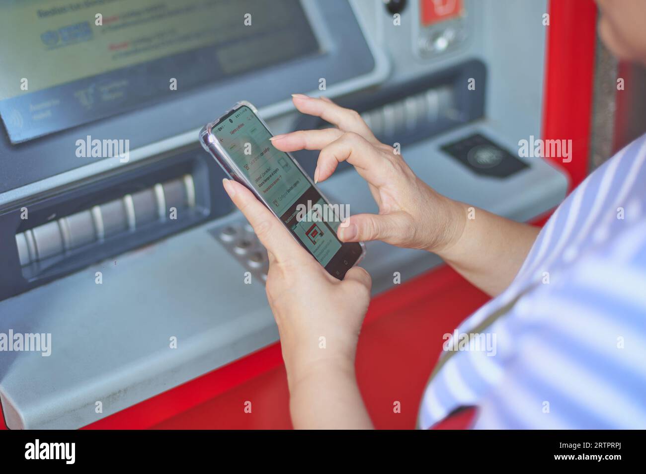 Femme méconnaissable tenant un téléphone portable devant un guichet automatique bancaire effectuant une opération bancaire en ligne. Banque D'Images