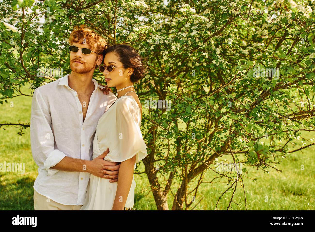 jeunes jeunes mariés multiethniques dans des lunettes de soleil et robe de mariée serrant dans le jardin vert, style boho Banque D'Images