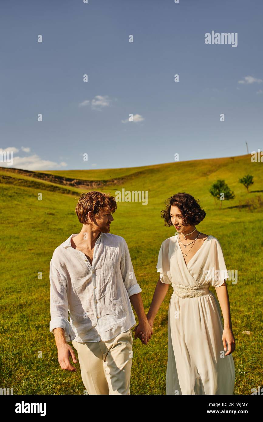 mariage à la campagne, couple multiethnique tenant la main dans le champ vert, juste mariés jeunes mariés Banque D'Images