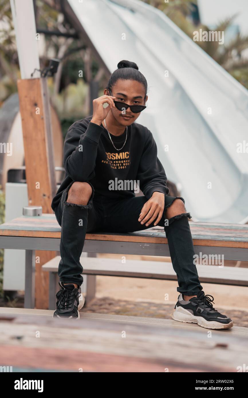 Un adolescent asiatique avec une chemise et des lunettes de soleil est assis sur un banc de parc très élégant et excentrique quand il rencontre ses amis dans un café Banque D'Images