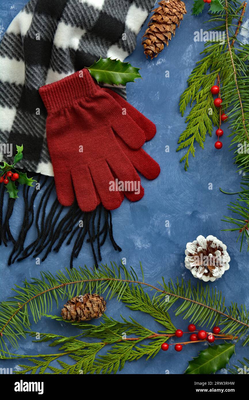 Écharpe frangée noire et blanche et gants rouges sur fond bleu avec verdure hivernale, baies et cônes dans la composition de pose plate. Concept hiver. Banque D'Images