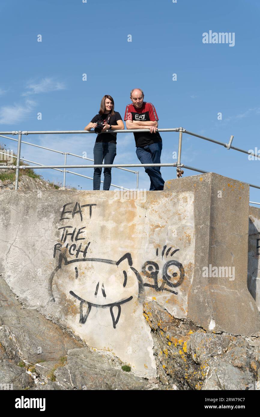 Deux personnes debout derrière des rampes au-dessus d'un mur de béton graffiné avec le message « mangez le riche » Banque D'Images