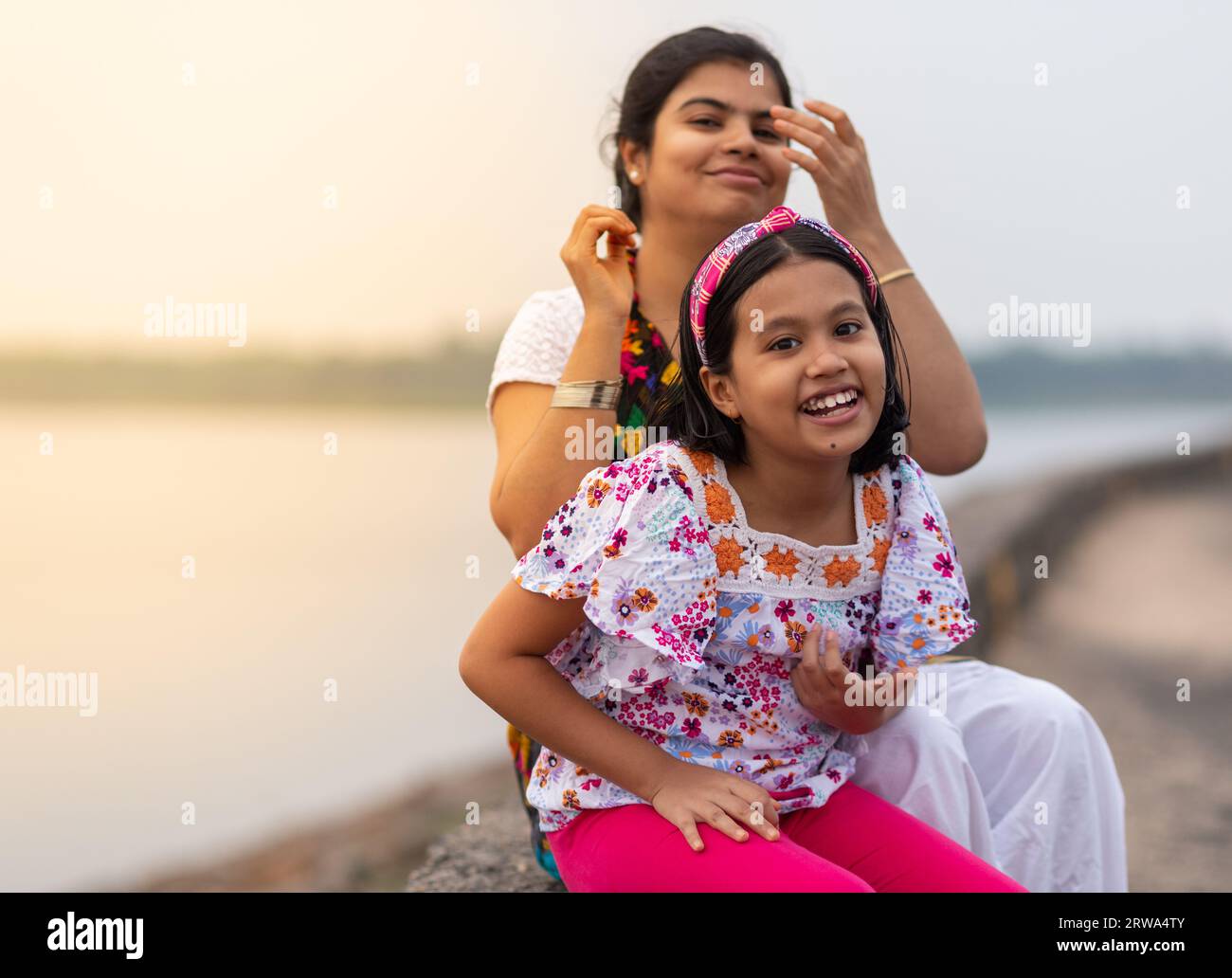 Mère et fille indiennes avec le visage souriant assis au bord de la rivière au coucher du soleil Banque D'Images