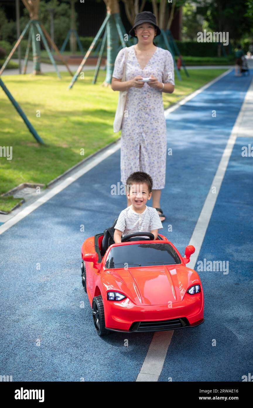 Mignon garçon en bas âge multiracial monte dans une voiture rouge télécommandée tandis que sa mère marche derrière lui conduisant un véhicule jouet Banque D'Images