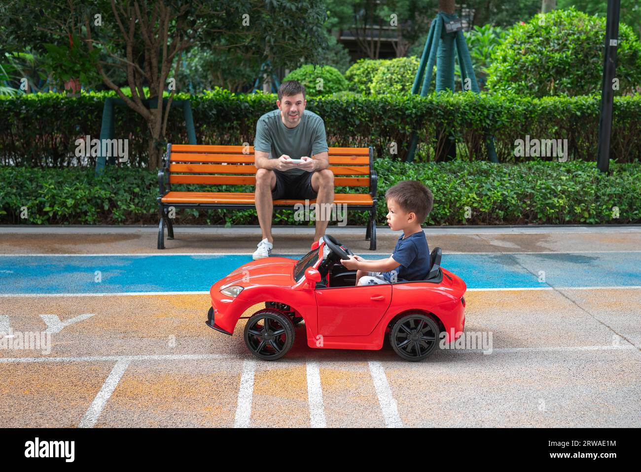 Mignon garçon en bas âge multiracial monte dans une voiture rouge télécommandée tandis que son père est assis sur le banc et conduit un véhicule jouet Banque D'Images